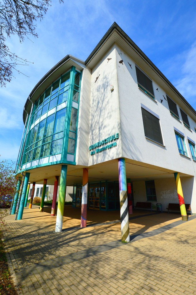 Das Foto zeigt den Haupteingang des Standortes Weißdornalle der Grundschule am Wasserturm. Vor der Eingangstür sind 14 bunt angemalte Säulen, die den überlappenden Vorbau stützen, der über den Eingang ragt.